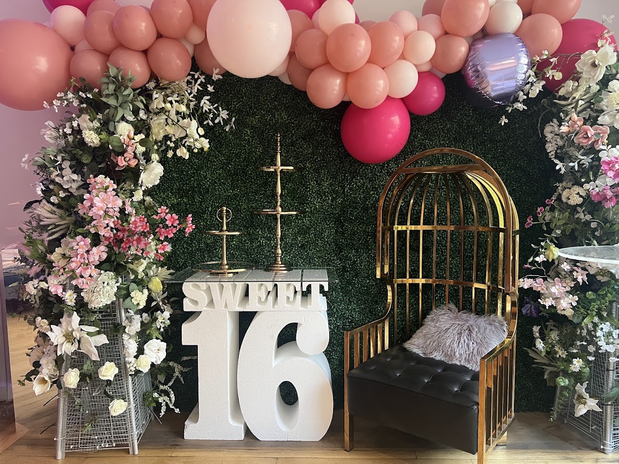 Sweet 16 backdrop with greenery wall, pink balloon garland, florals, and a gold cage chair