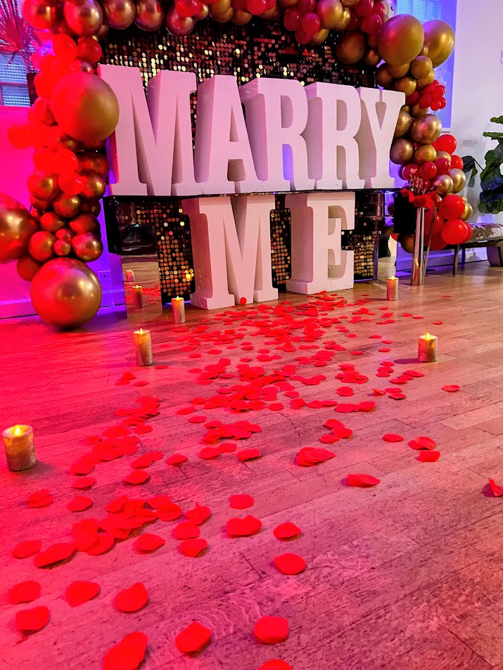 Marry Me marquee letters with red and gold balloons, candles, and rose petals on the floor